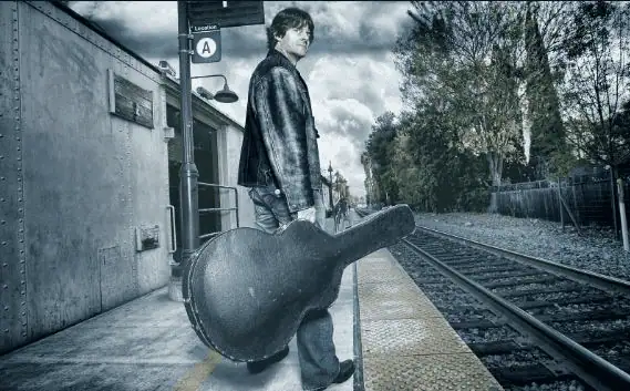 Musician standing on a train platform holding a guitar case beside empty railroad tracks.