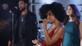 Female vocalist singing passionately onstage with backup singers behind her.