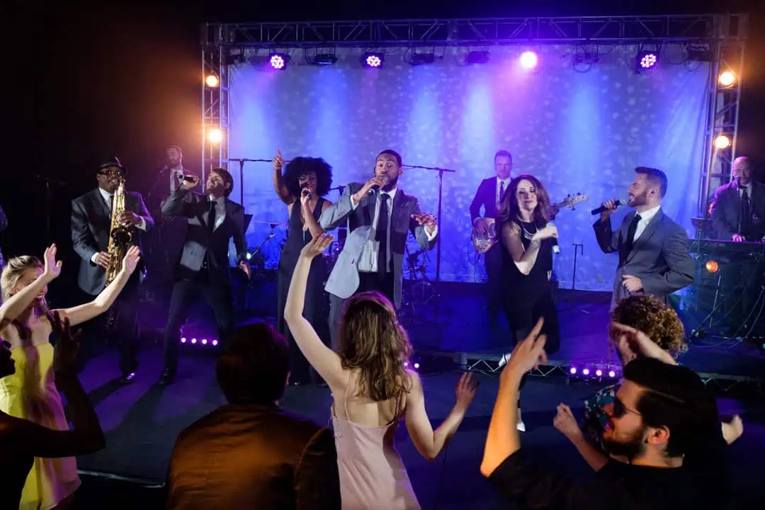 Live band performing onstage with multiple vocalists, musicians, and an enthusiastic crowd dancing in front.
