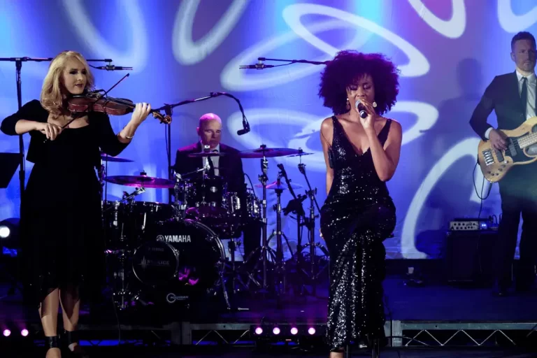 Singer, violinist, drummer, and bassist performing together on a stage with blue lighting.