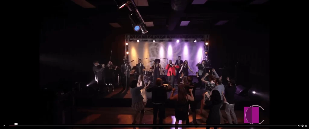 Live band performing onstage with vocalists, musicians, and a crowd dancing in front under dramatic stage lighting.
