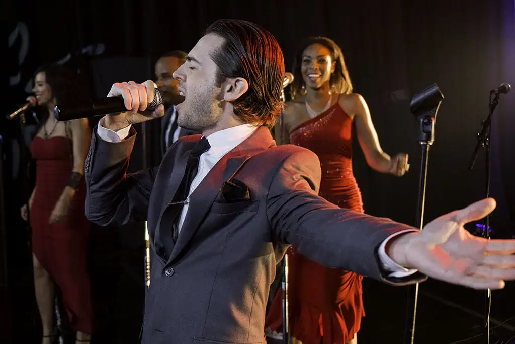 Male vocalist performing passionately onstage with female backup singers in red dresses.