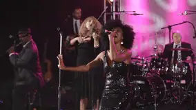Female vocalist performing energetically onstage with musicians and a violinist behind her.