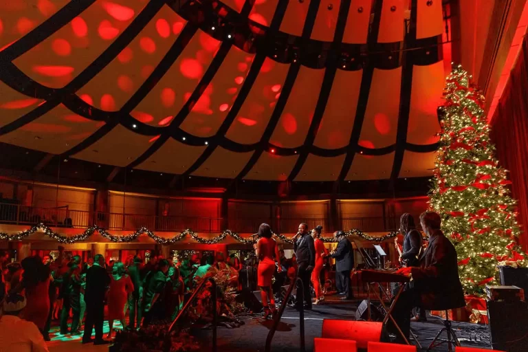A holiday gala with a live band performing on stage, a large decorated Christmas tree, festive red and gold lighting, and guests dancing in an elegant venue.