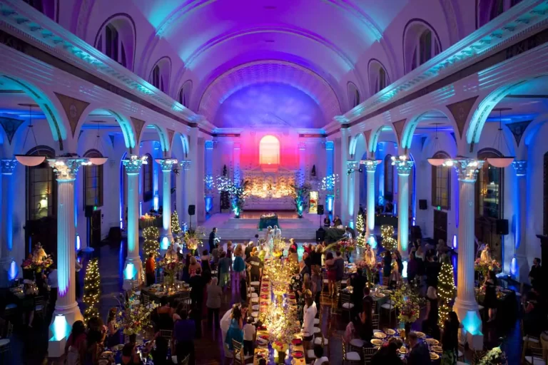 A grand ballroom event with colorful blue and purple uplighting, tall columns, decorated tables, and guests celebrating in an elegant venue.
