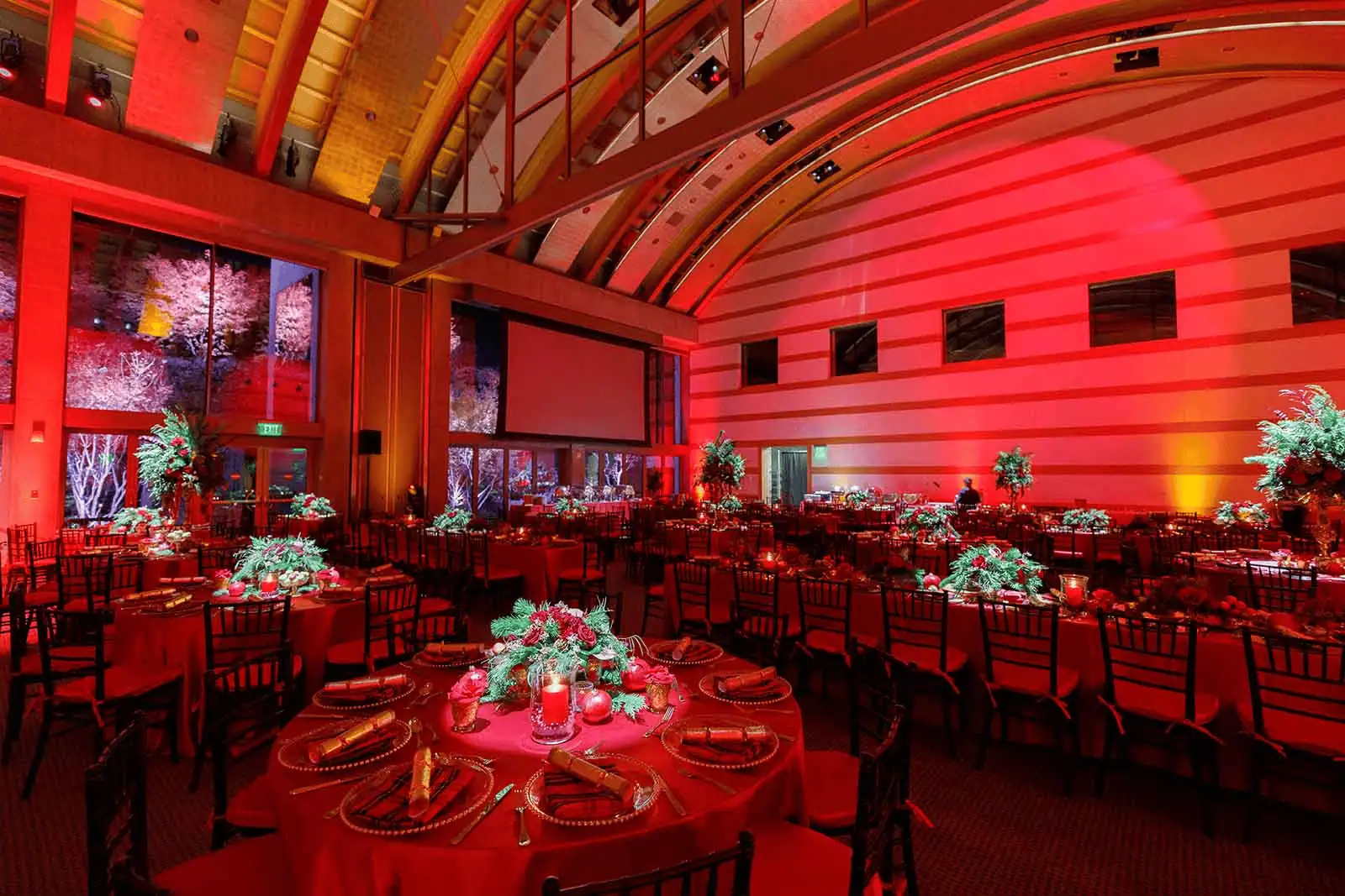 A festive event space decorated in red lighting with round tables, floral centerpieces, and elegant holiday-inspired décor.