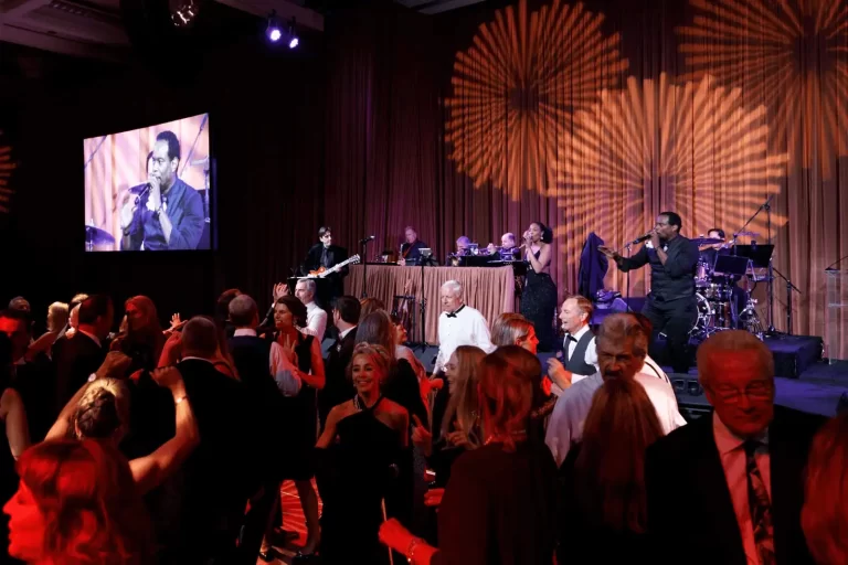 Guests dancing in a ballroom while a live band performs on stage with warm patterned lighting and a large video screen.