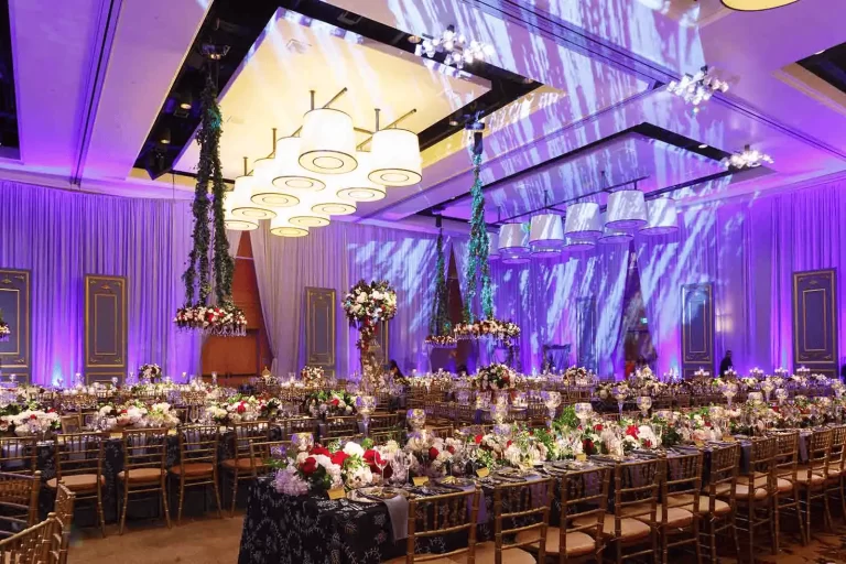 Lavish ballroom decorated with floral centerpieces, gold seating, and dramatic purple event lighting.