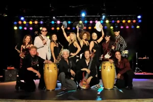 Large party band posing onstage with vocalists, musicians, percussionists, and colorful stage lighting.