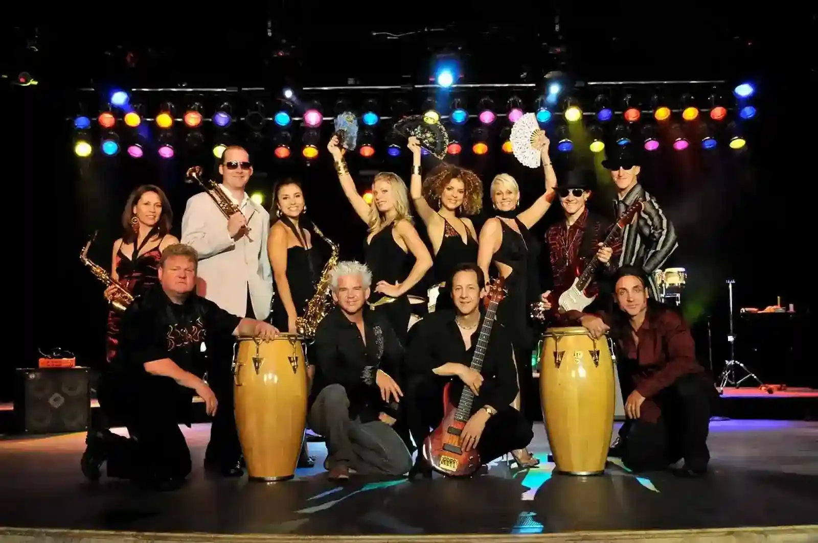 Large party band posing onstage with vocalists, musicians, percussionists, and colorful stage lighting.