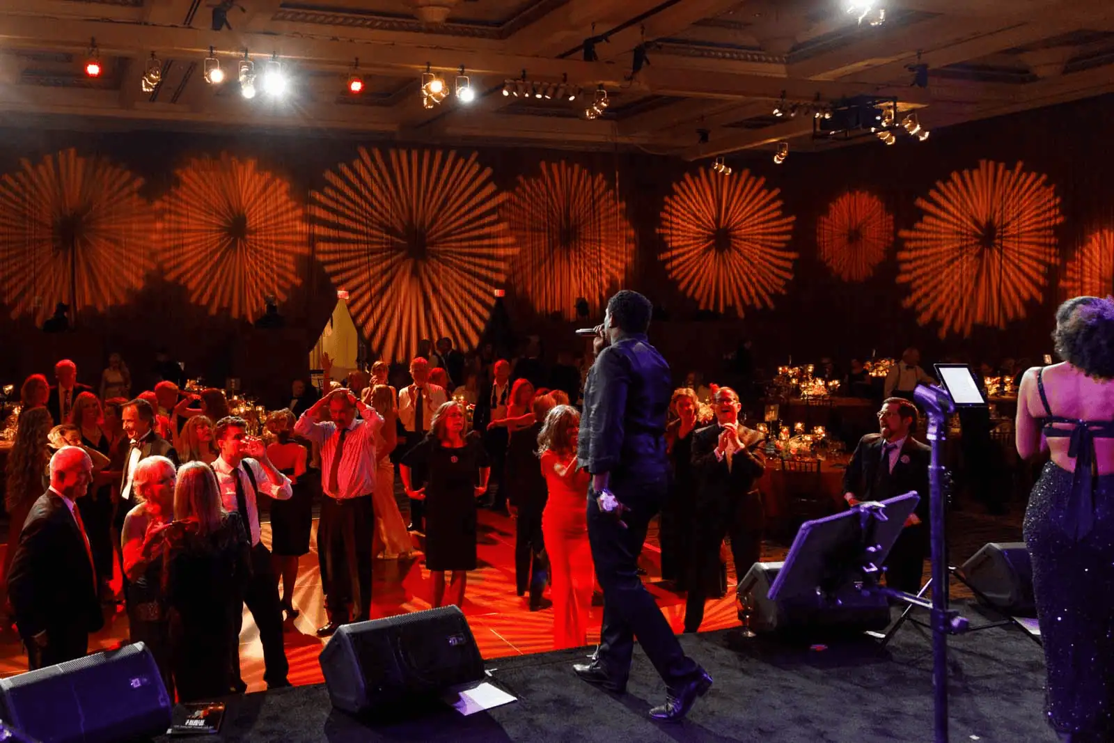 Live performers on stage energizing a ballroom crowd as warm patterned lighting fills the room during an evening celebration.